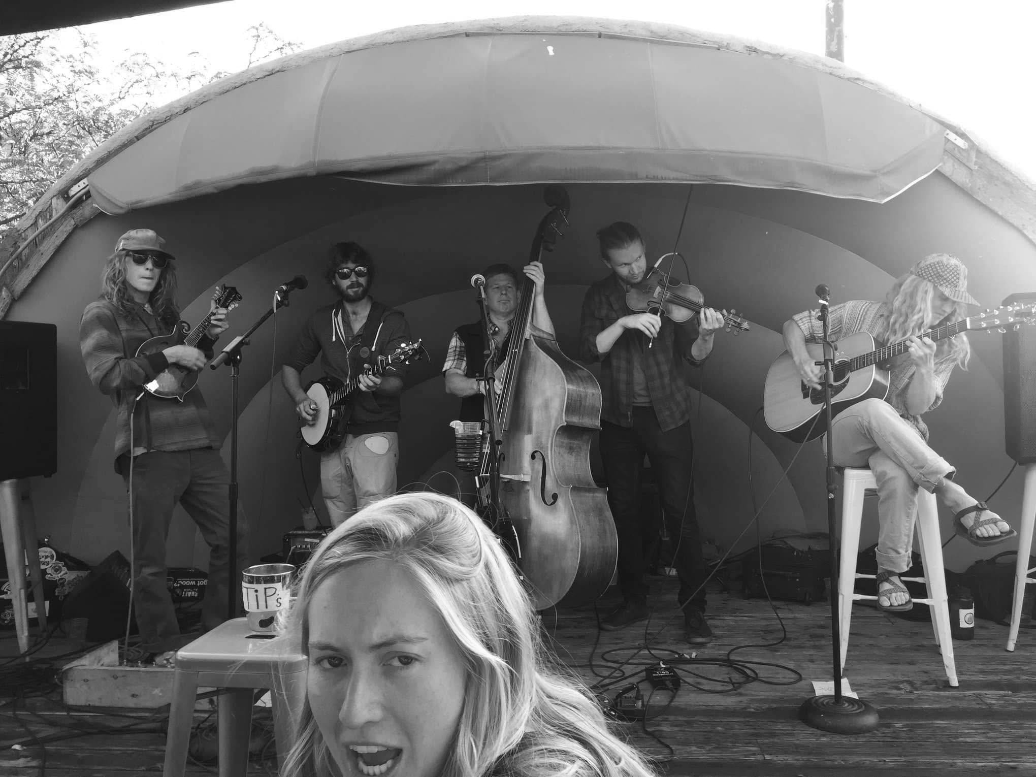 Five-piece string band performing under a dome stage with a tip jar on a stool in front